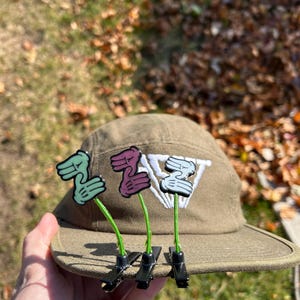 May include: A khaki-colored cap with three decorative hand-shaped pins. The pins are green, purple, and white, attached with green stems and black clips. The cap has a white emblem.
