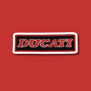 Peut inclure: Écusson brodé avec le mot "DUCATI" en rouge, souligné de blanc, sur un fond rectangulaire noir. L'écusson a une bordure blanche et est posé sur un fond rouge. Idéal pour les passionnés de motos.