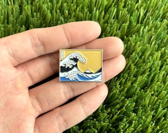 May include: A silver enamel pin featuring a blue and white wave with a yellow background. The wave is a depiction of the famous Japanese artwork "The Great Wave off Kanagawa".