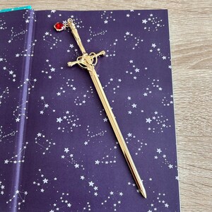 May include: A gold sword bookmark with a red gem on the handle. The bookmark is lying on a purple book cover with a white constellation pattern.