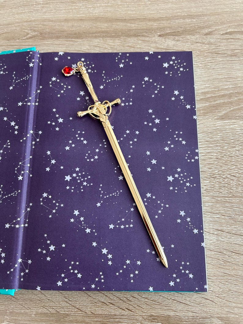 May include: A gold sword bookmark with a red gem on the handle. The bookmark is lying on a purple book cover with a white constellation pattern.