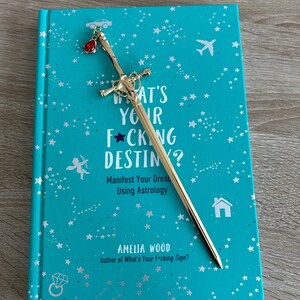 May include: A turquoise blue book with a gold sword bookmark. The book cover features a star pattern and the text "What's Your F*cking Destiny? Manifest Your Dreams Using Astrology" and "Amelia Wood, Author of What's Your F*cking Sign?"