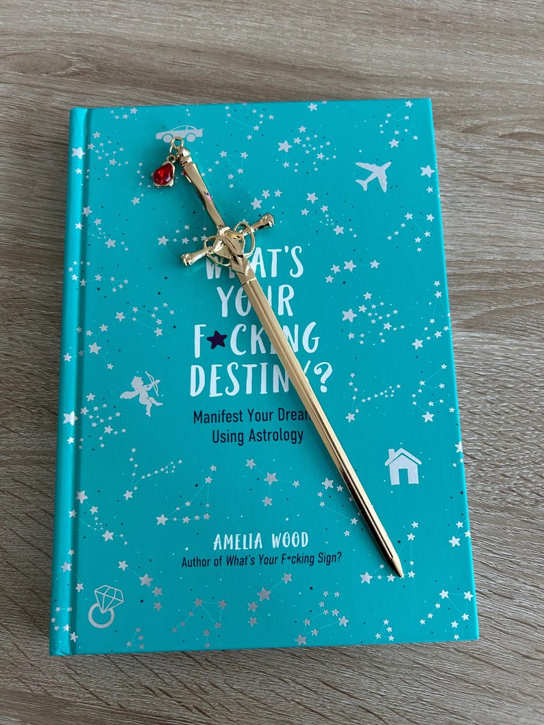 May include: A turquoise blue book with a gold sword bookmark. The book cover features a star pattern and the text "What's Your F*cking Destiny? Manifest Your Dreams Using Astrology" and "Amelia Wood, Author of What's Your F*cking Sign?"