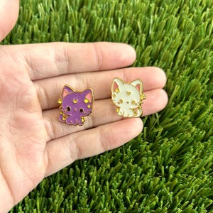 May include: Two enamel pins featuring a cartoon cat design. The cat on the left is purple with gold stars and the cat on the right is white with gold stars.