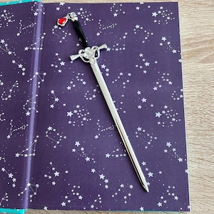 May include: A silver sword bookmark with a red gem on the handle. The bookmark is laying on a book with a dark purple cover and a white constellation pattern.