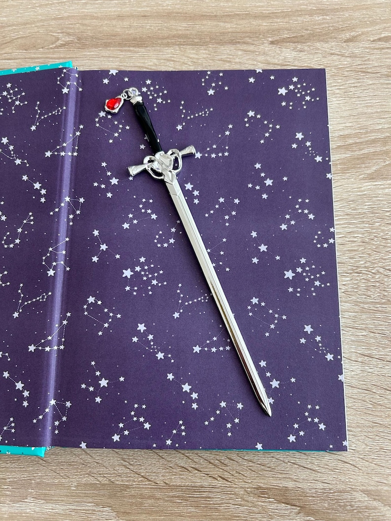 May include: A silver sword bookmark with a red gem on the handle. The bookmark is laying on a book with a dark purple cover and a white constellation pattern.