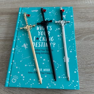 May include: Three decorative sword-shaped bookmarks with red heart-shaped accents. The bookmarks are gold, black, and silver. The bookmarks are on top of a teal book with a star pattern and the text "What's Your F*cking Destiny?" The book's author is Amia Wood.