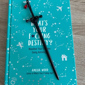 May include: A teal blue book with silver stars and constellations printed on the cover. The book title is "What's Your F*cking Destiny?" with the subtitle "Manifest Your Dreams Using Astrology." The author is Amelia Wood, author of "What's Your F*cking Sign?" A black sword bookmark with a red gem is on top of the book.