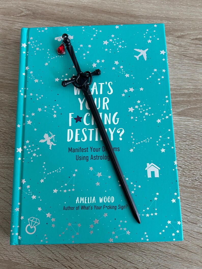 May include: A teal blue book with silver stars and constellations printed on the cover. The book title is "What's Your F*cking Destiny?" with the subtitle "Manifest Your Dreams Using Astrology." The author is Amelia Wood, author of "What's Your F*cking Sign?" A black sword bookmark with a red gem is on top of the book.