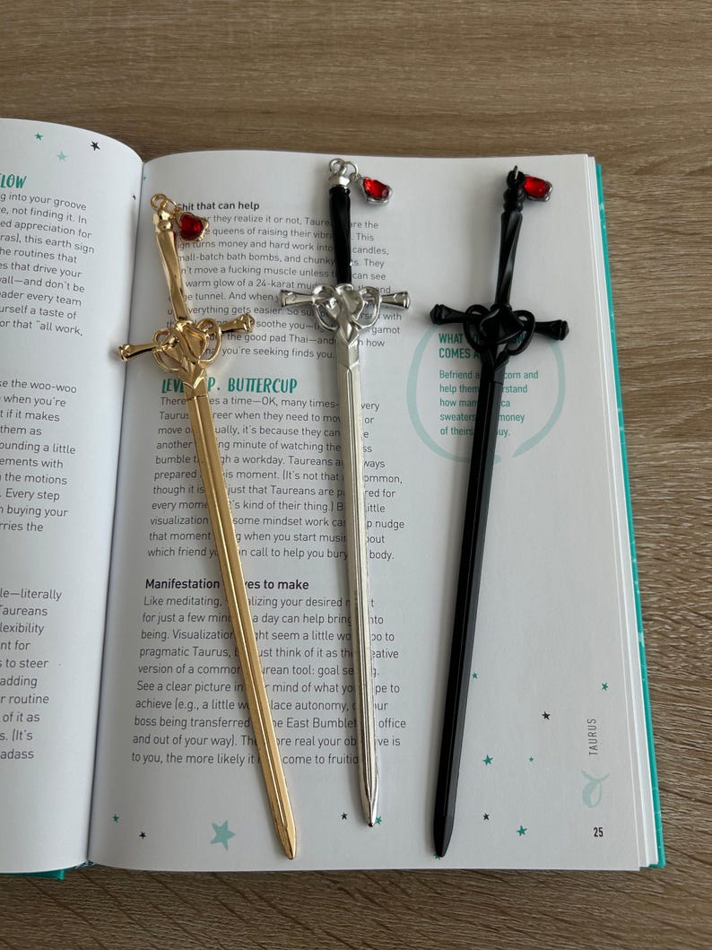 May include: Three decorative metal bookmarks in the shape of swords with red gem accents. The bookmarks are gold, silver, and black. The text "WHAT COMES AROUND" is visible on the black bookmark.