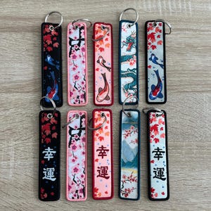May include: Ten colorful keychains with different designs. The keychains feature images of koi fish, cherry blossoms, dragons, and Japanese characters. The keychains are all black and white with accents of pink, blue, and green.