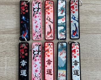 Könnte beinhalten: Zehn bunte Schlüsselanhänger mit verschiedenen Designs. Die Schlüsselanhänger zeigen Bilder von Koi-Karpfen, Kirschblüten, Drachen und japanischen Schriftzeichen. Die Schlüsselanhänger sind alle schwarz-weiß mit Akzenten in Pink, Blau und Grün.