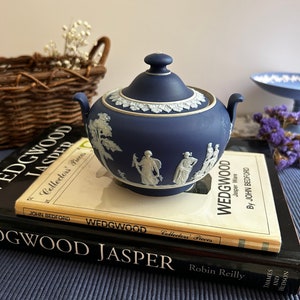 May include: A blue and white Wedgwood Jasperware sugar bowl with a lid. The bowl is decorated with a white cameo scene of figures in a landscape. The bowl is sitting on top of a stack of books about Wedgwood.
