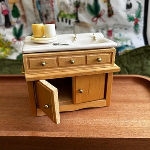 May include: A miniature wooden cabinet with a white porcelain sink and two small cups on a plate. The cabinet has three drawers and a door that is open. The cabinet is made of light wood with gold-colored hardware.
