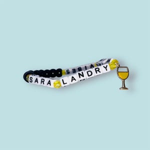 May include: A beaded bracelet spelling out "SARA LANDRY" in white letter beads. The bracelet features black and yellow beads, and a gold-coloured wine glass charm. The words "GOOD VIBES" are also on the bracelet.