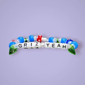 Puede incluir: Una pulsera de cuentas con letras blancas cuadradas que forman la palabra GRIZ YEAH, intercaladas con cuentas azules, transparentes y con forma de hongo rojo. El diseño incluye dos dijes verdes en forma de hoja en los extremos del accesorio.