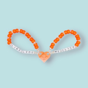May include: Orange beaded friendship bracelet with a heart-shaped Lego charm. The bracelet has the words "Like Lego" and "We Click" spelled out on it.