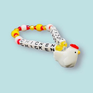 May include: A colourful beaded bracelet spelling "CHICKEN" in white letter beads. The bracelet includes yellow, red, and white beads, and a white chicken charm with a red comb and yellow beak.