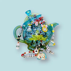 May include: A blue teapot filled with colourful beads and charms, including a white rabbit, a playing card, and a teacup. The teapot is decorated with the words "Beyond Wonderland".