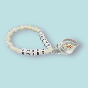May include: White beaded bracelet with the name "EMBER" spelled out in black letters on the beads. The bracelet has a clear seashell charm with a small seashell inside.