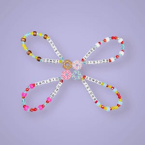 May include: A decorative beaded design in a butterfly shape. It features four loops with the words "DONUT DISTURB" spelled out in white beads. The loops are decorated with colourful beads and miniature donut charms in various colours.