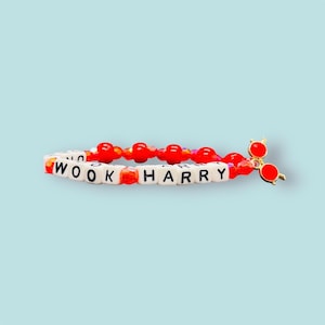 May include: A beaded bracelet with white letter beads spelling "WOOK HARRY" and red beads. A gold-coloured charm of round red glasses is attached. The bracelet is on a light blue background.