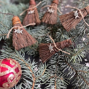 May include: Miniature brown brooms with silver triple moon charms, tied with twine, rest on a bed of evergreen branches. A red and gold ornament is in the lower left corner. The brooms are likely decorative items.