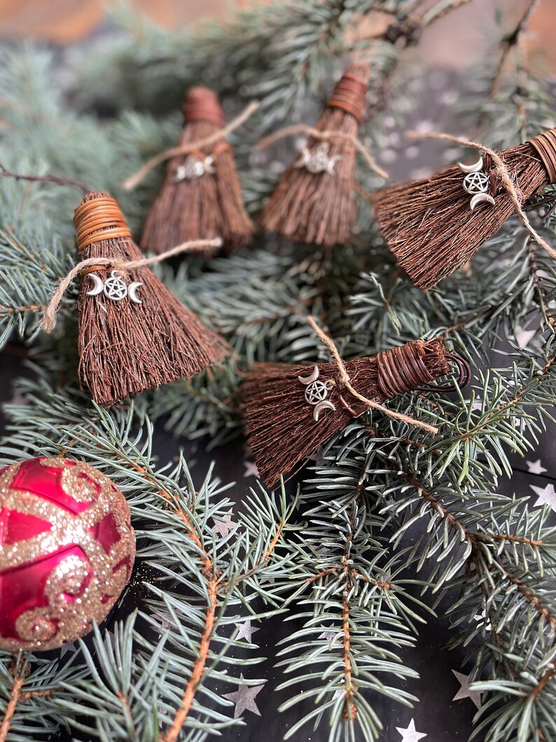 May include: Miniature brown brooms with silver triple moon charms, tied with twine, rest on a bed of evergreen branches. A red and gold ornament is in the lower left corner. The brooms are likely decorative items.