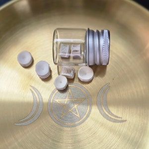 May include: A small glass bottle with a silver lid containing white ceramic pieces. The bottle is sitting on a gold metal tray with a pentagram and crescent moon design.