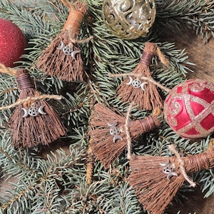 May include: A collection of small, brown broom ornaments with silver crescent moon and pentacle charms, tied with twine. The brooms are arranged on a bed of evergreen branches, with red and gold Christmas ornaments.