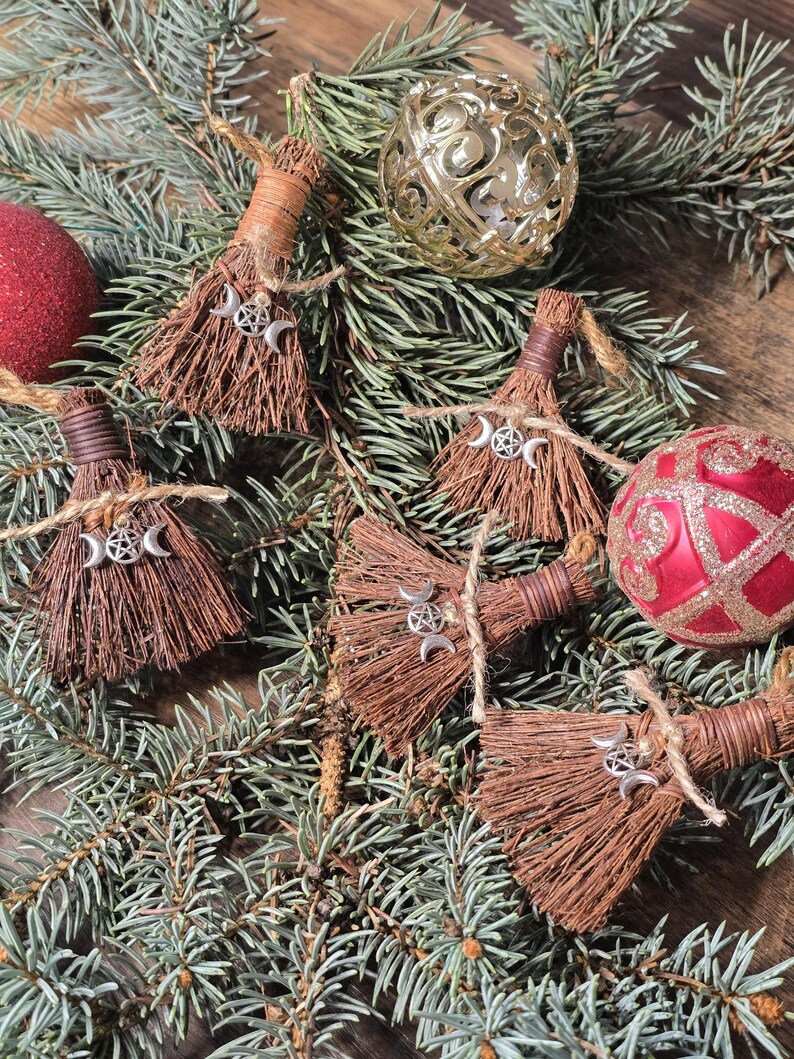 May include: A collection of small, brown broom ornaments with silver crescent moon and pentacle charms, tied with twine. The brooms are arranged on a bed of evergreen branches, with red and gold Christmas ornaments.
