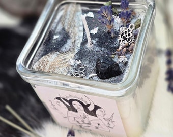 May include: A square glass candle jar with a black and white design, a black stone, and dried lavender flowers. The jar has a label with a black and white illustration of three women with crescent moons above their heads.