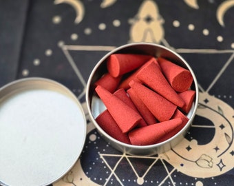 Könnte beinhalten: Eine silberne Dose gefüllt mit roten Räucherkegeln. Die Dose steht auf einer schwarzen und goldenen gemusterten Oberfläche.