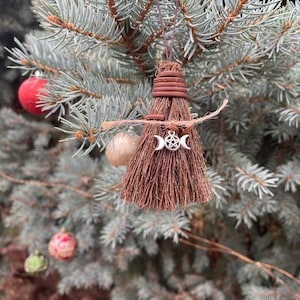 May include: A small, brown broom ornament with a silver triple moon and pentacle charm, hanging from a blue-green evergreen branch. The broom is tied with brown twine and has a rustic, natural appearance. Christmas ornaments are visible in the background.