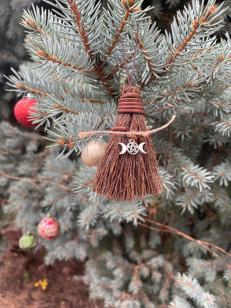 May include: A small, brown broom ornament with a silver triple moon and pentacle charm, hanging from a blue-green evergreen branch. The broom is tied with brown twine and has a rustic, natural appearance. Christmas ornaments are visible in the background.