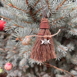 May include: A small, handmade broom ornament with a brown straw brush and a wrapped handle. A silver charm with a triple moon and pentacle hangs from the broom. The ornament is suspended from a brown wire and twine, displayed on a blue-green evergreen branch.