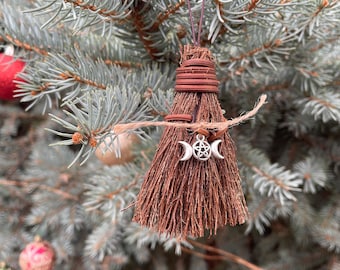 May include: A miniature broom ornament with a brown straw brush and a wrapped handle. A silver charm with a triple moon and pentacle hangs from the broom. The ornament is suspended from a brown wire and twine, displayed on a blue-green evergreen branch.