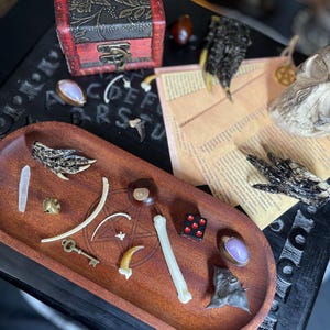 May include: An oval wooden tray featuring an engraved pentagram, containing a collection of small items including a white bone, a brass key, a red die with black dots, a purple stone, a brown nut, a claw, and a small metal bell.