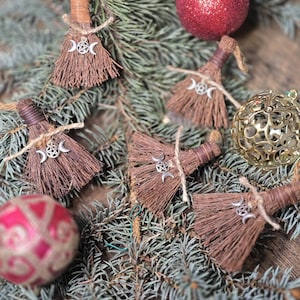 May include: A collection of brown broom ornaments with silver triple moon charms, tied with twine, displayed on a bed of evergreen branches. Red and gold Christmas ornaments are also visible, creating a festive holiday scene.