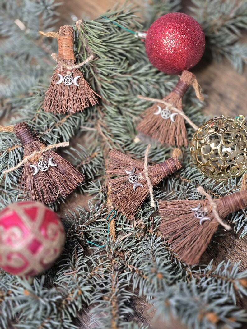 May include: A collection of brown broom ornaments with silver triple moon charms, tied with twine, displayed on a bed of evergreen branches. Red and gold Christmas ornaments are also visible, creating a festive holiday scene.