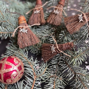 May include: Miniature brown broom ornaments with silver triple moon charms, tied with twine. The brooms are arranged on a bed of evergreen branches, with a red and gold glittery Christmas ornament in the lower left corner. The background is a dark surface with white stars.