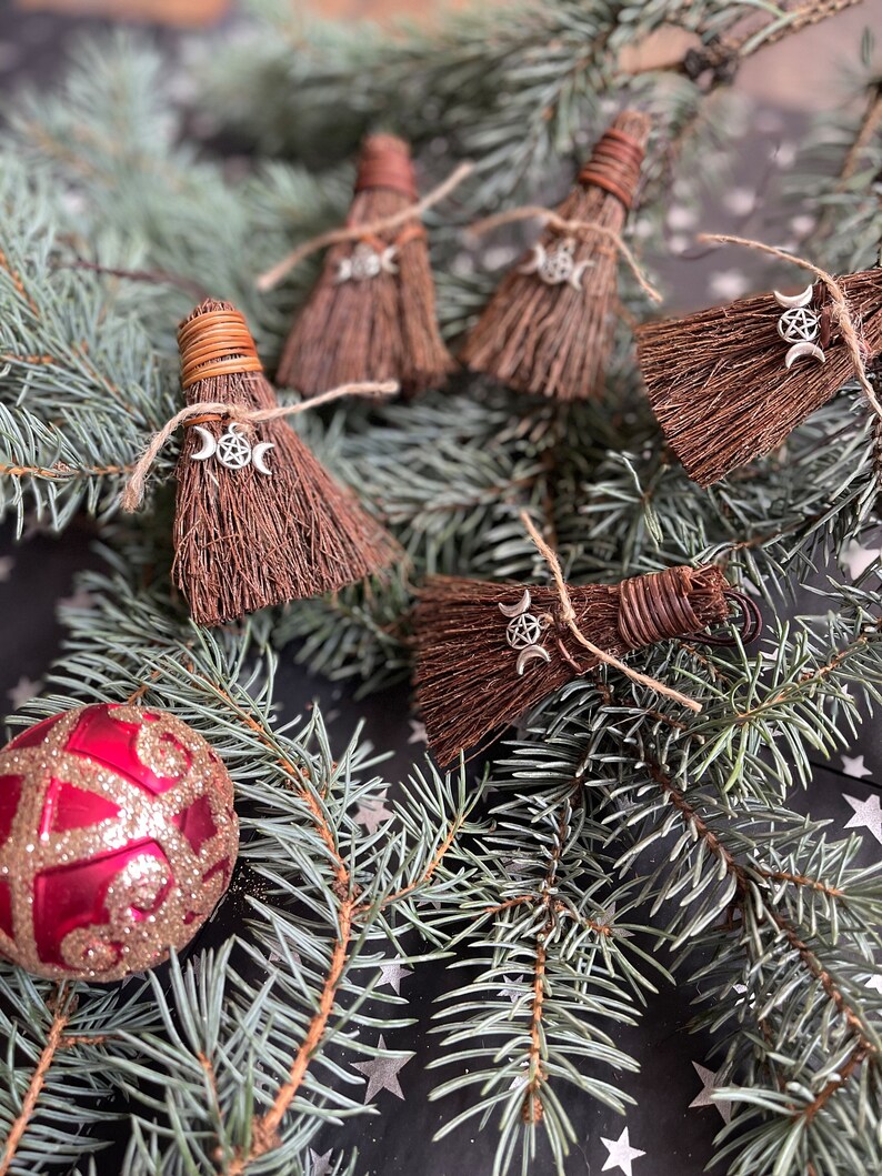 May include: Miniature brown broom ornaments with silver triple moon charms, tied with twine. The brooms are arranged on a bed of evergreen branches, with a red and gold glittery Christmas ornament in the lower left corner. The background is a dark surface with white stars.