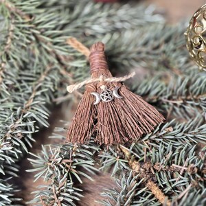 May include: A miniature broom ornament made of brown bristles and twine, adorned with a silver charm featuring a crescent moon and star design. The broom is set against a backdrop of evergreen branches and a red ornament.