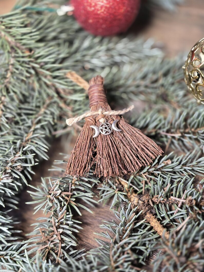 May include: A miniature broom ornament made of brown bristles and twine, adorned with a silver charm featuring a crescent moon and star design. The broom is set against a backdrop of evergreen branches and a red ornament.