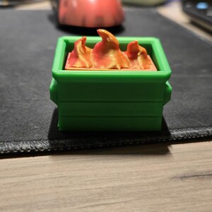 May include: A green miniature dumpster with orange and gold flames inside. The dumpster is rectangular with a hinged lid and a textured surface. The flames have a textured, layered appearance.