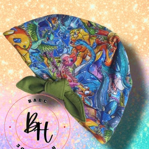 May include: A baby bonnet with a vibrant dinosaur print and a green bow. The bonnet showcases a variety of cartoon dinosaurs in blue, yellow, orange, and green. The text "BABE HANDMADE BH" is in a circle.
