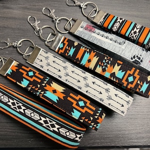May include: Six colorful keychains with different geometric patterns, including arrows, diamonds, and stripes. The keychains are made of fabric and have silver metal key rings.