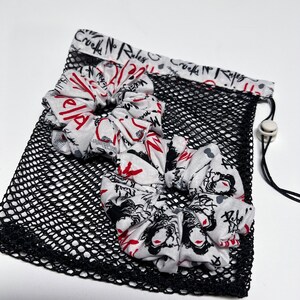 May include: Two white fabric scrunchies with a black and red print featuring the text "Cruella No Rules". The scrunchies are in a black mesh bag with a drawstring.