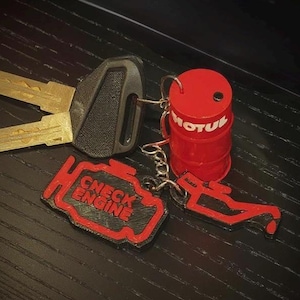 May include: A set of keys with a black key fob, alongside a red Motul barrel keyring and two other red and black keyrings with engine-related designs. One keyring reads "CHECK ENGINE".