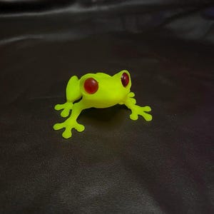 May include: A small, lime green frog ornament with bright red eyes. The frog is posed with its legs spread, as if about to leap. The background is a dark, textured surface.